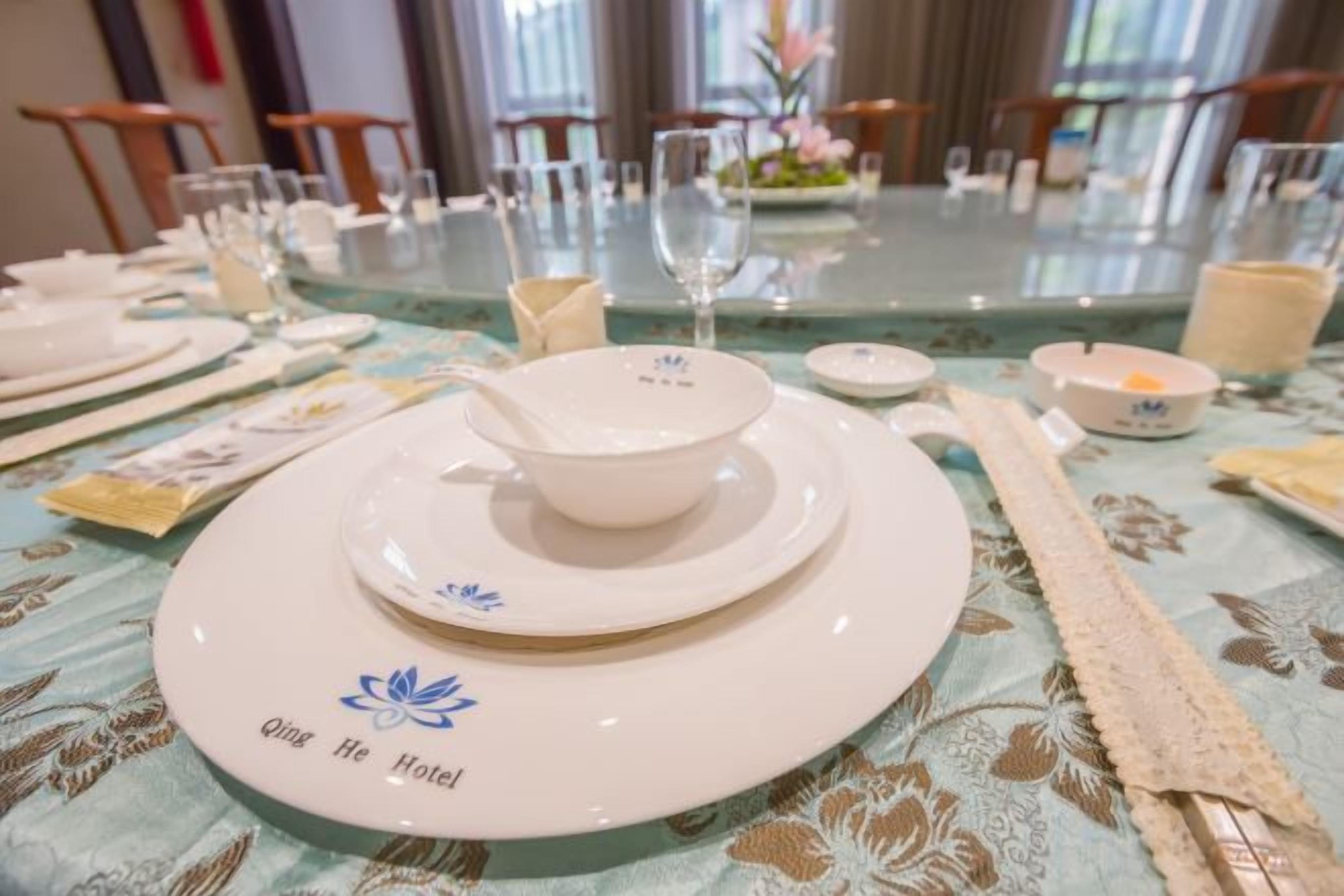 A dining table set with plates, bowls, and cutlery, with a floral tablecloth and a 'Qing He Hotel' logo.