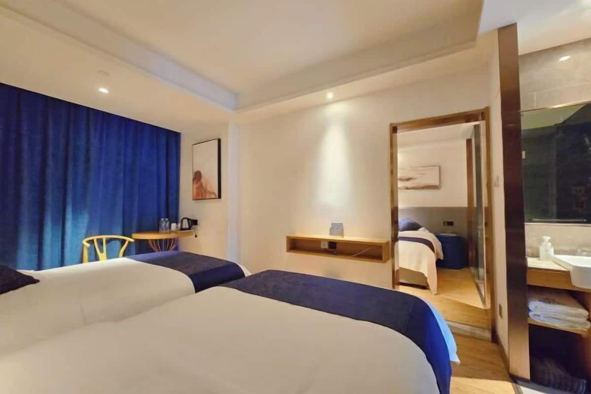 Habitación de hotel con dos camas, un escritorio, una silla, una mesita, un espejo y un baño con lavamanos y perchero para toallas.