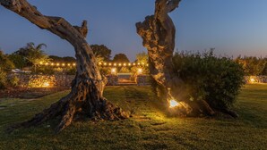A tree-lined pathway leading to a stone wall with a gate, surrounded by a well-lit garden at dusk. - Tenuta Del Leccino - Torre Vado (Torre vado)