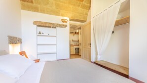 A bedroom with a large bed, a wooden bench, a stone wall, and a kitchen in the background. - Tenuta Del Leccino - Torre Vado (Torre vado)