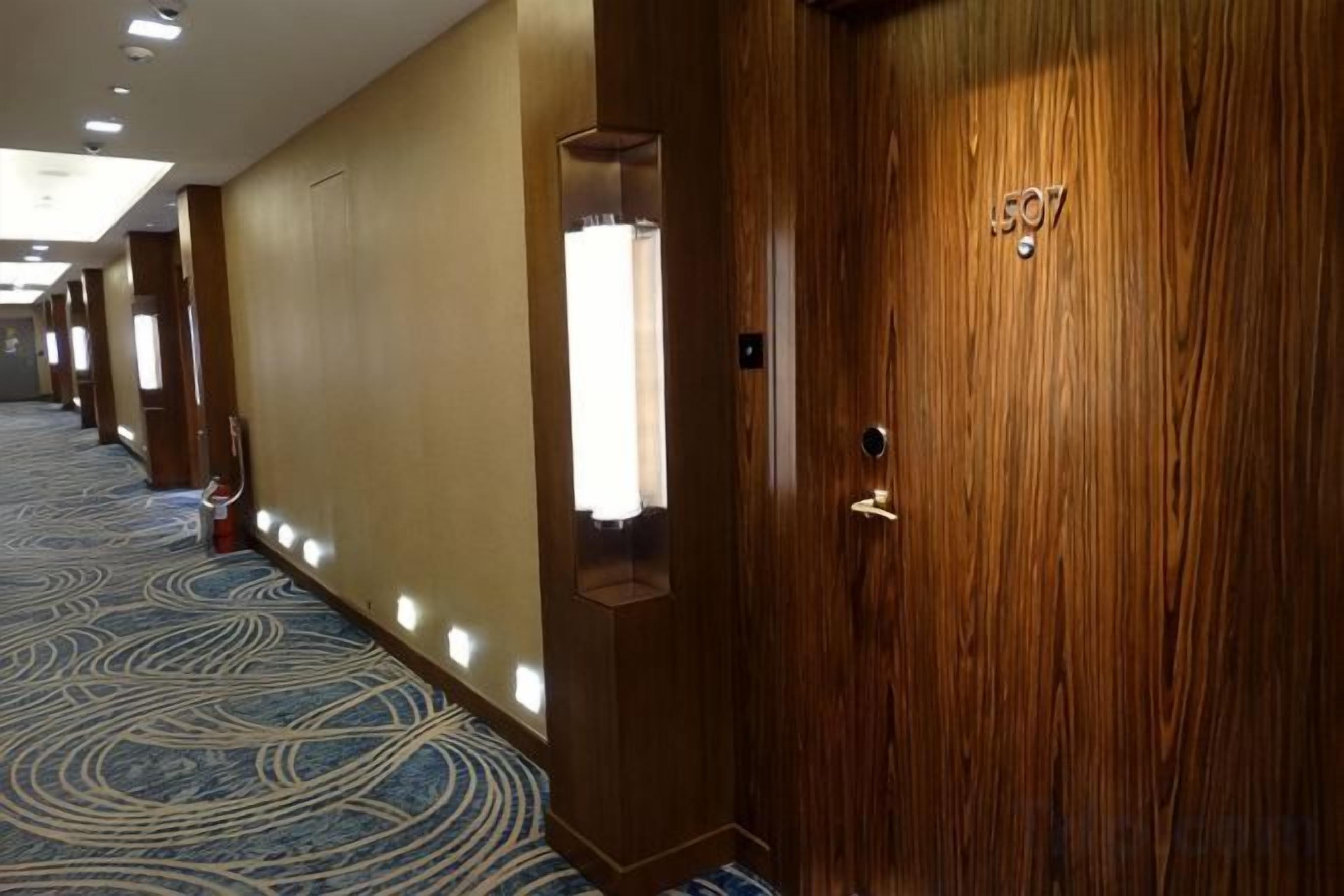 Corredor de hotel com portas de madeira, carpete estampado e iluminação embutida.