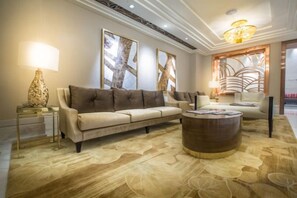 Un salon spacieux avec un canapé beige, une table basse ronde et un grand tapis. On y trouve deux lampes fixées au mur et un miroir décoratif.