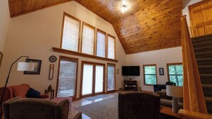Ein geräumiges Wohnzimmer mit hohen Decken, Holzbalken, großen Fenstern, einem Flachbildfernseher und einer Treppe, die in einen oberen Bereich führt.