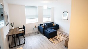 A modern living room with a dark blue sofa, a wooden dining table with chairs, and a small side table with a bottle and glasses.