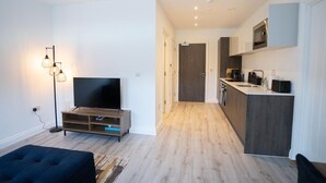 A modern apartment with a living room featuring a flat-screen TV, a dark blue sofa, and a floor lamp. The kitchen has a dark wood countertop, a microwave, and a coffee machine.