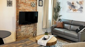 Een moderne woonkamer met een bakstenen muur, een flatscreen televisie, een grijze bank en een houten eettafel.