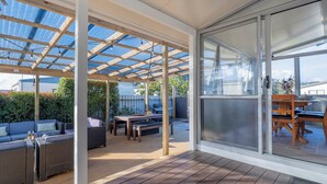 Un patio techado al aire libre con piso de madera, un área de estar con sofá y mesa, y una puerta de vidrio que da a vistas del exterior.