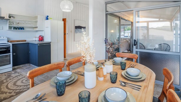 Una cocina moderna con armarios blancos, un comedor con una mesa redonda dispuesta para cuatro personas, y vistas al exterior a través de una puerta de cristal.