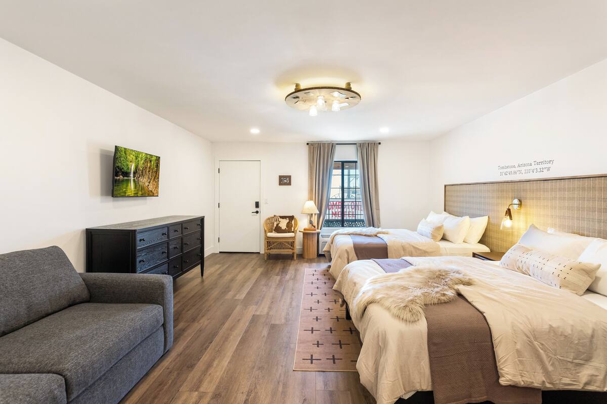 Ein modernes Schlafzimmer mit Holzboden, einer Couch, zwei Betten mit beige farbenen Bettwäsche, einem Fernseher an der Wand und einem Fenster mit Vorhängen.