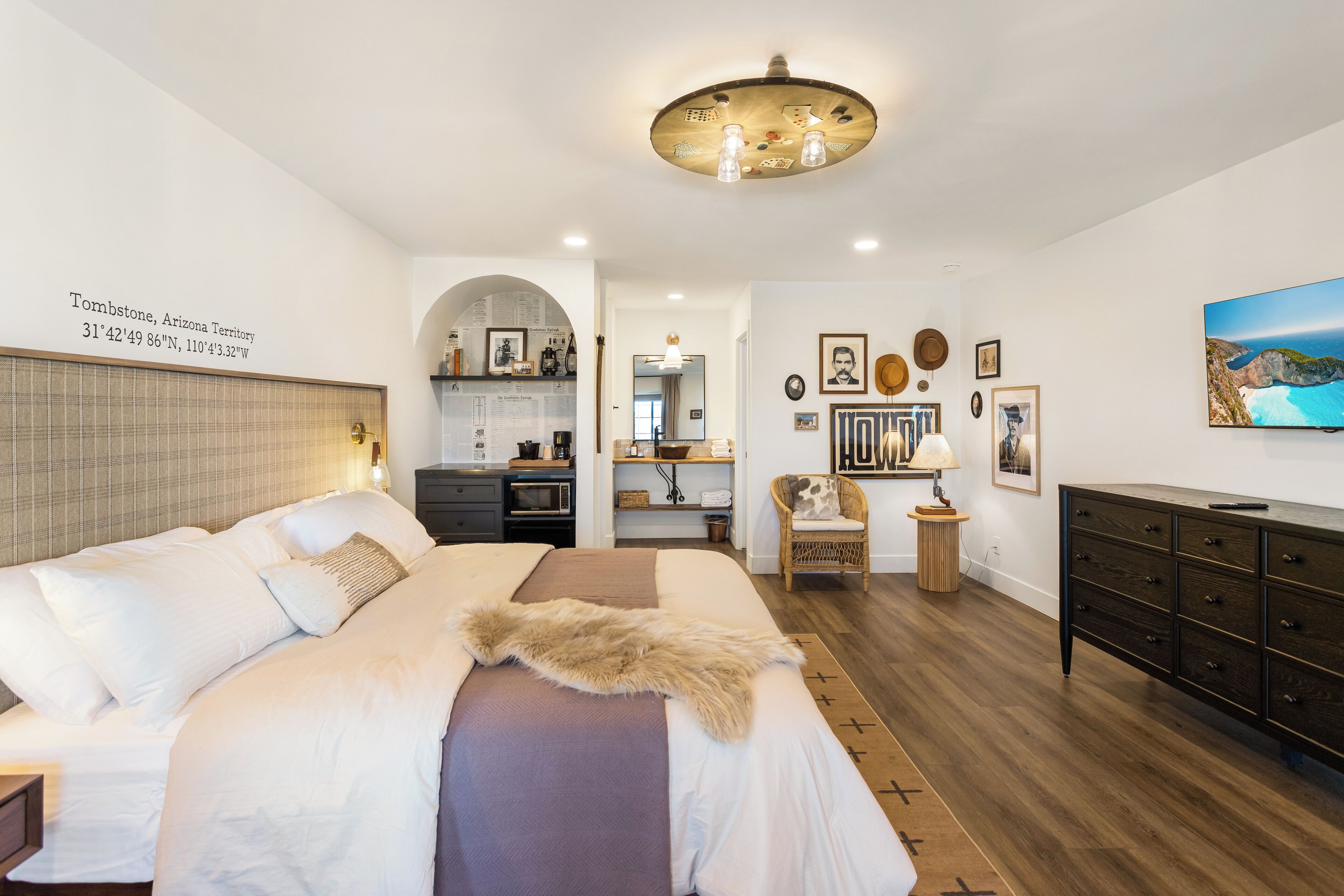 A bedroom with a large bed, a wooden dresser, a chair, and a dining area with a kitchen in the background.