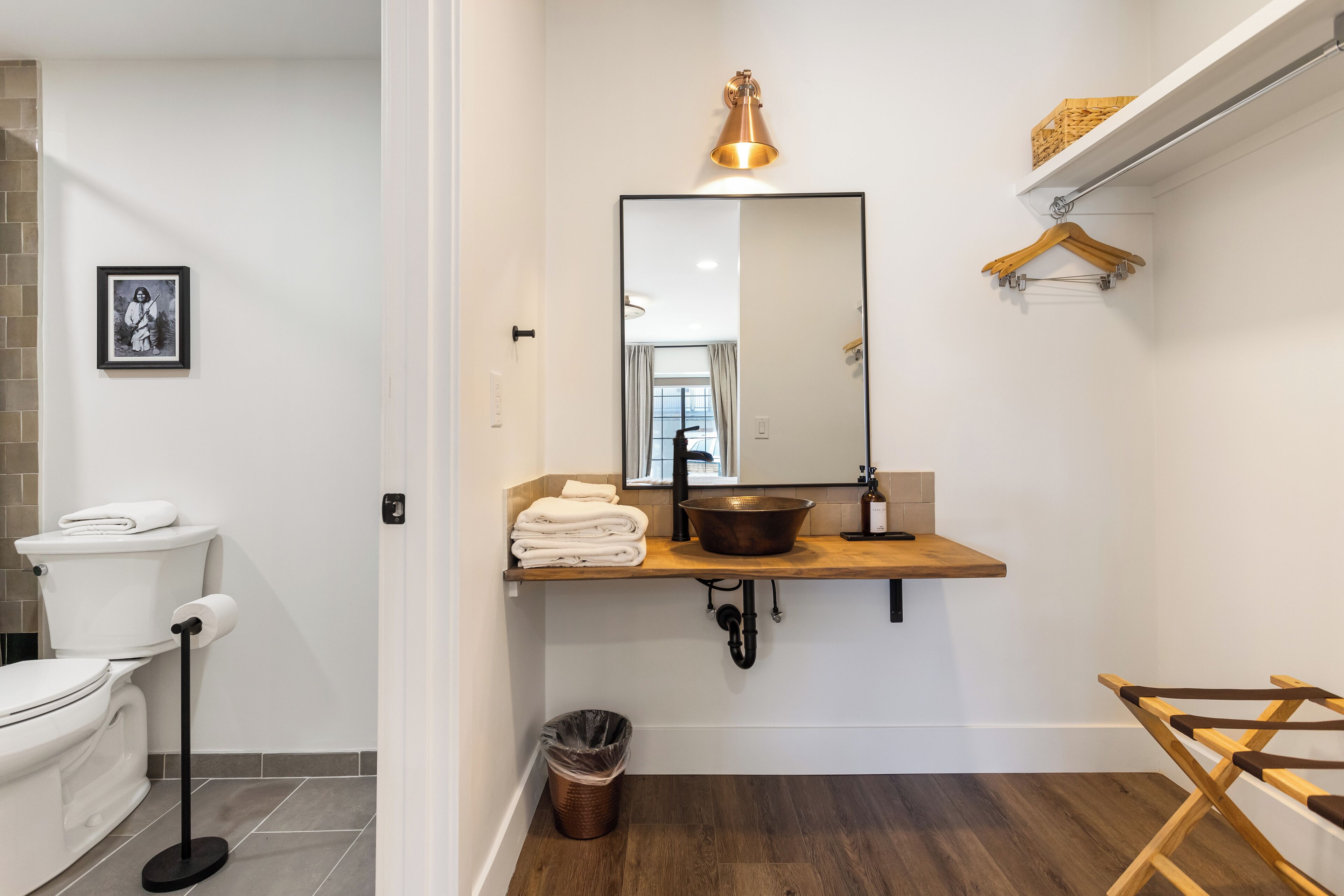 A modern bathroom with a white toilet, a sink with a copper basin, a mirror, and a wooden shelf with a towel rack.