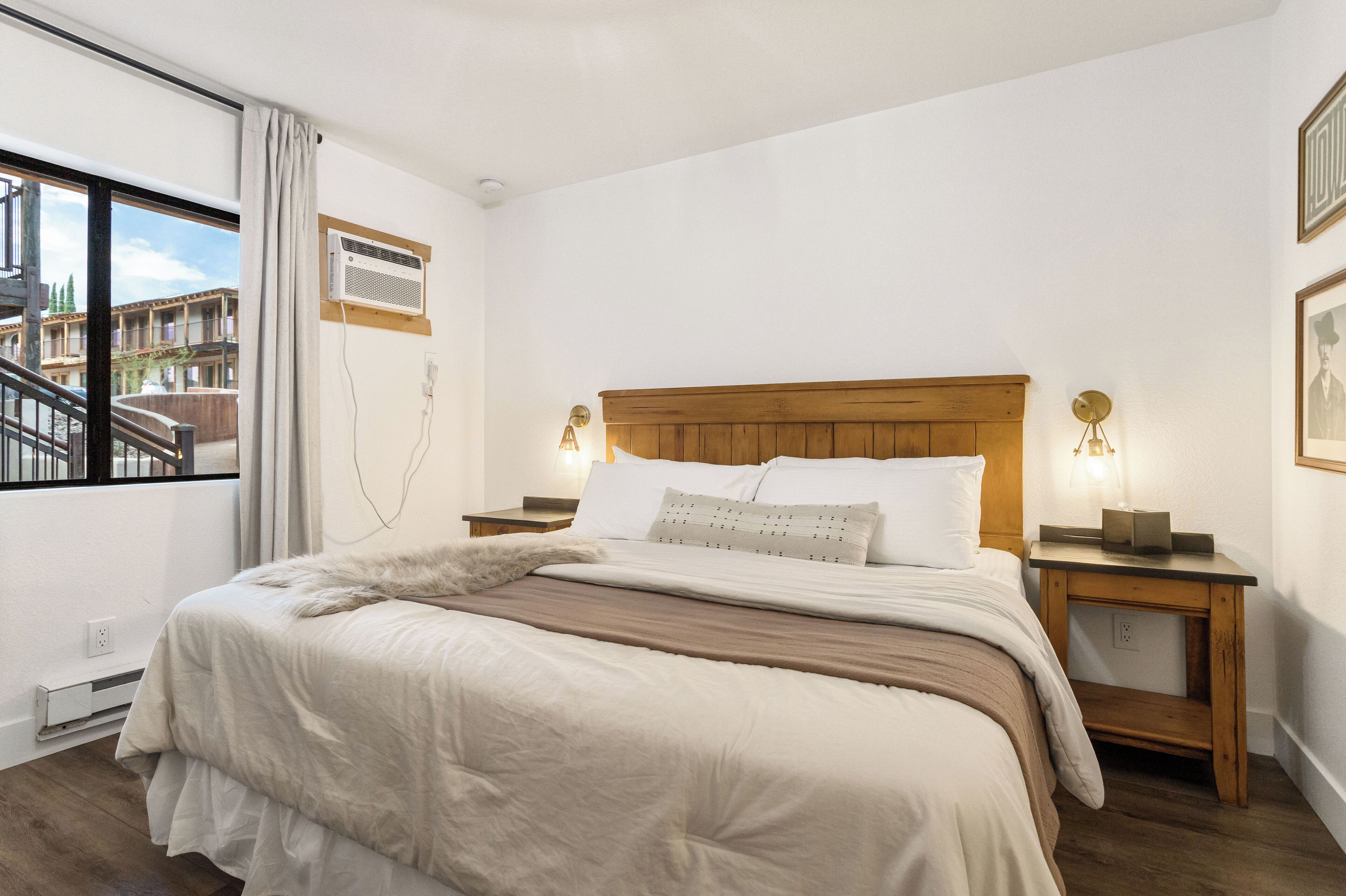 A bedroom with a large bed, wooden headboard, two bedside tables with lamps, a window with a view of buildings, and a wall-mounted air conditioner.