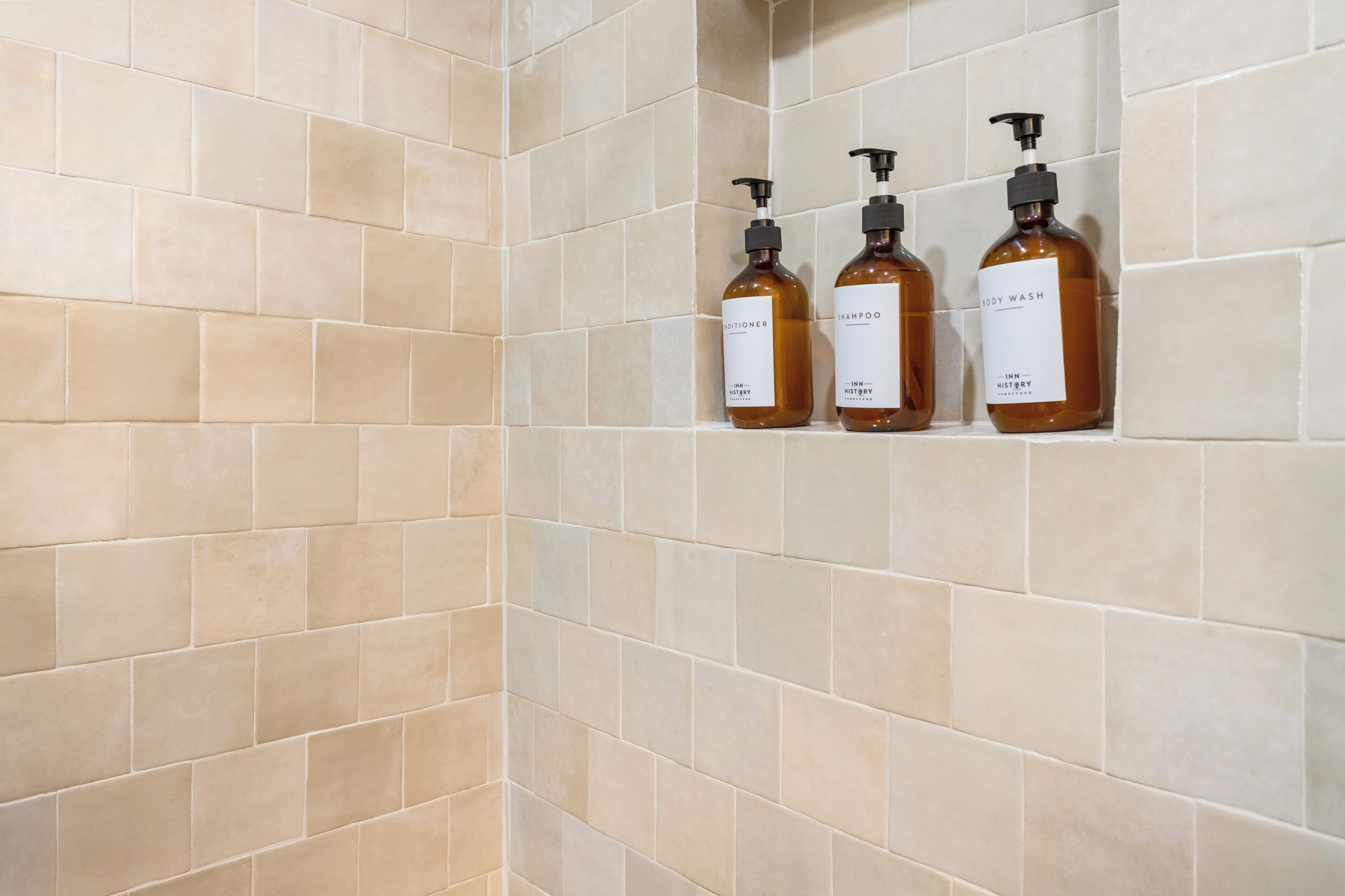 A bathroom with beige tiles and three bottles of body wash and shampoo on the wall.