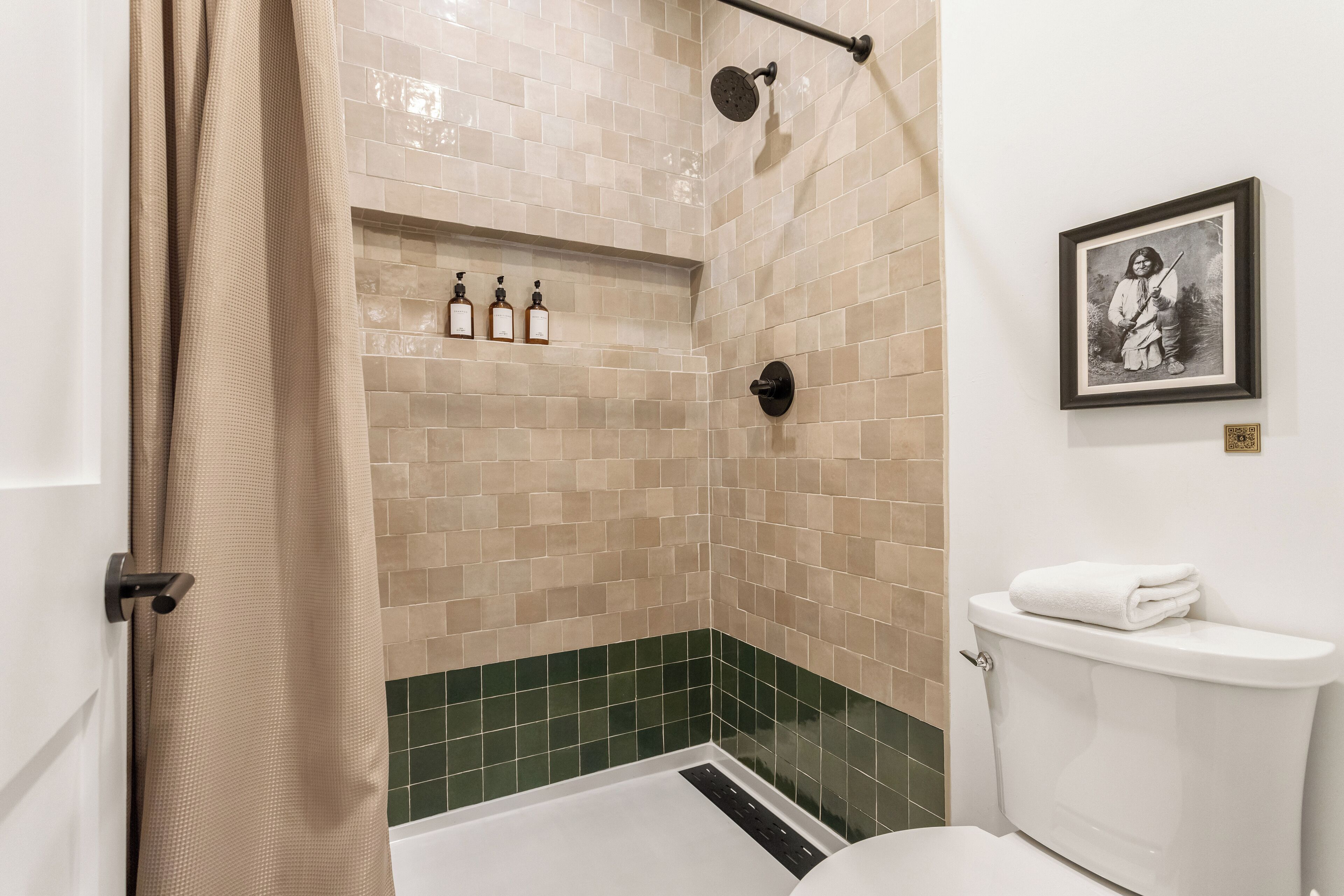 A bathroom with beige and green tiled walls, a shower with a handheld showerhead, a toilet with a towel on top, and a framed picture on the wall.