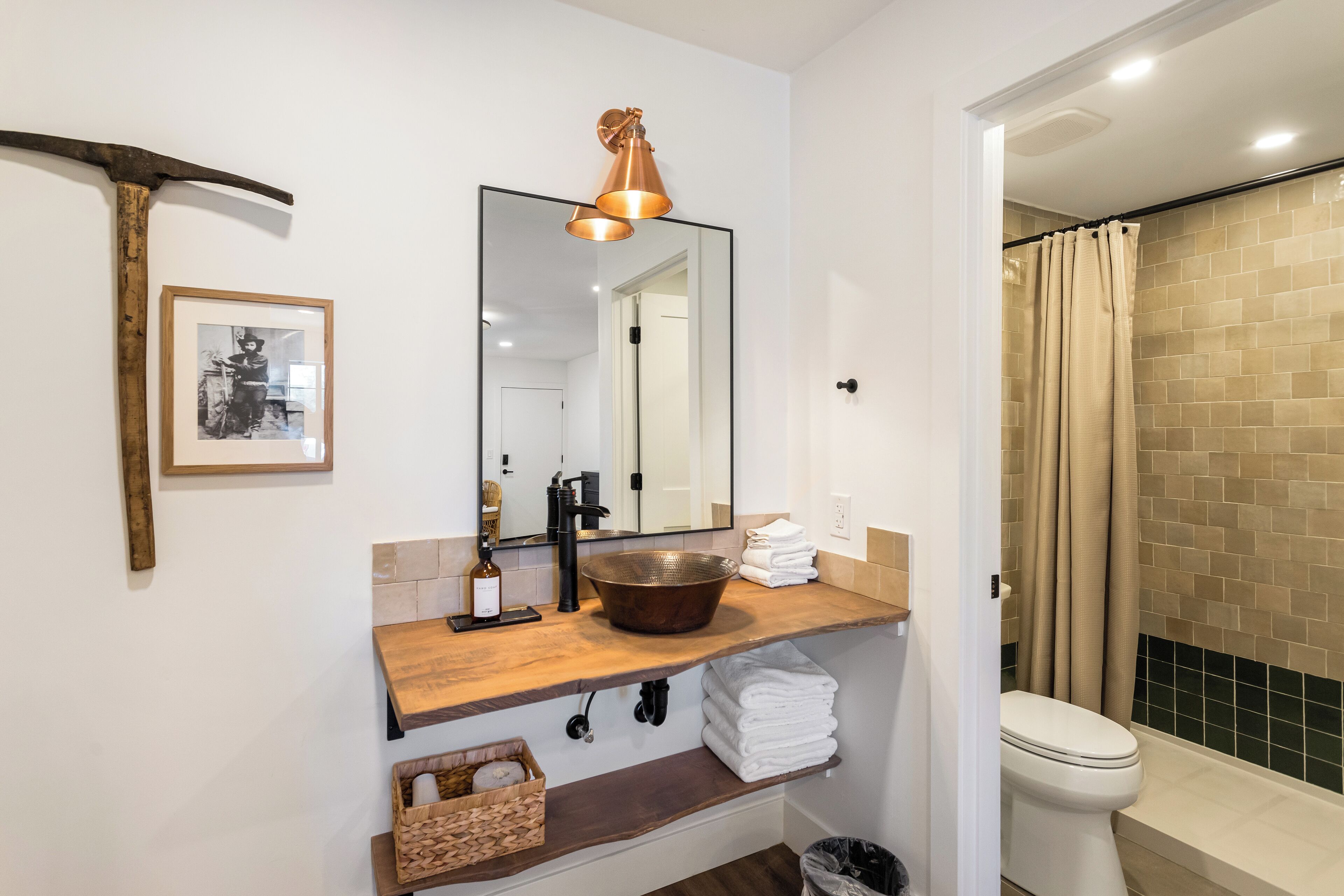 Een badkamer met een houten wastafelmeubel, een grote spiegel, een koperen lamp, een ingelijst kunstwerk en een douche met beige tegels.