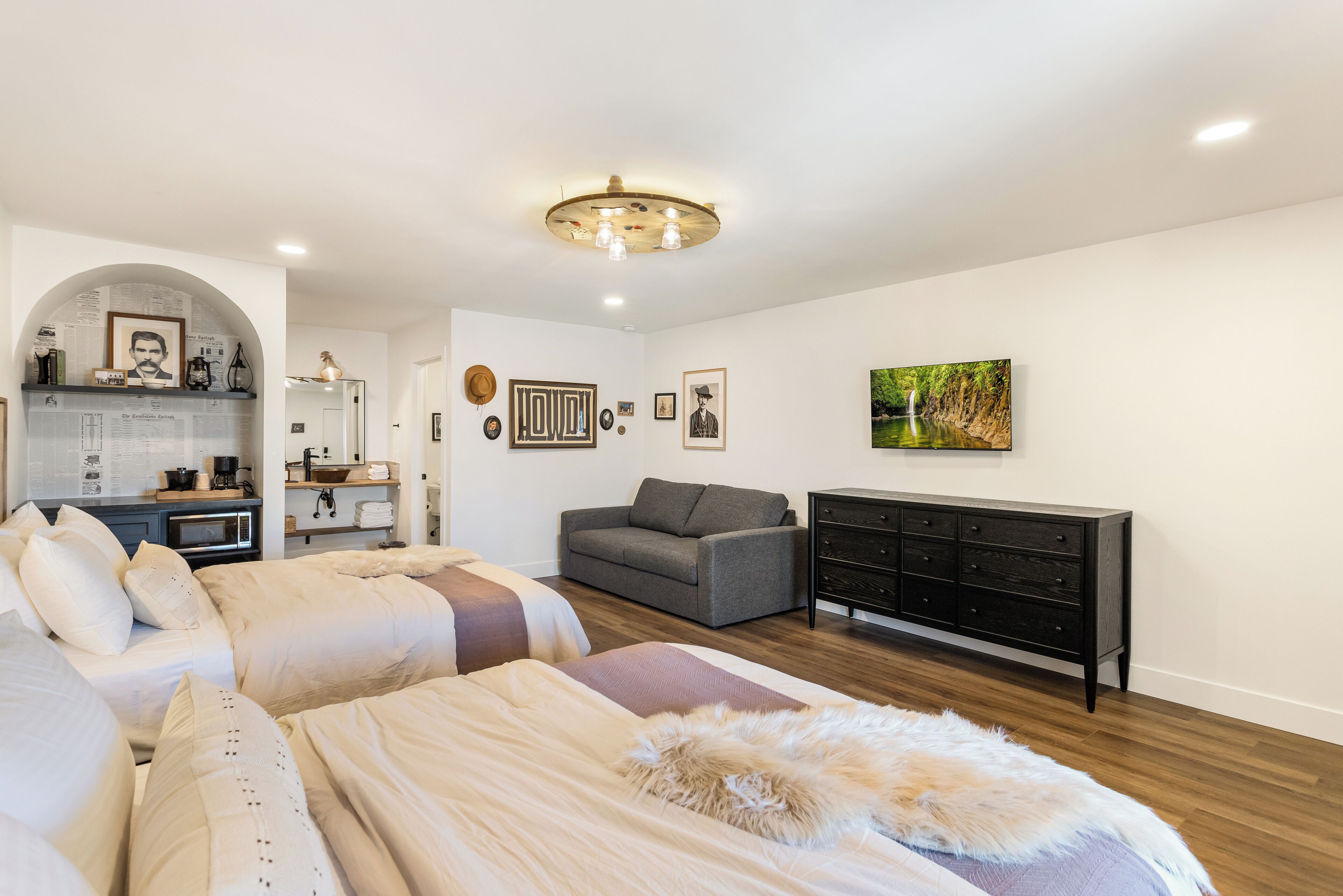 A modern bedroom with a large bed, a sofa, a dresser, and a kitchen area in the background.