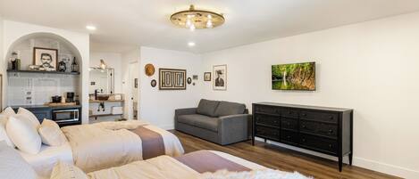 A modern bedroom with a large bed, a sofa, a dresser, and a kitchen area in the background.