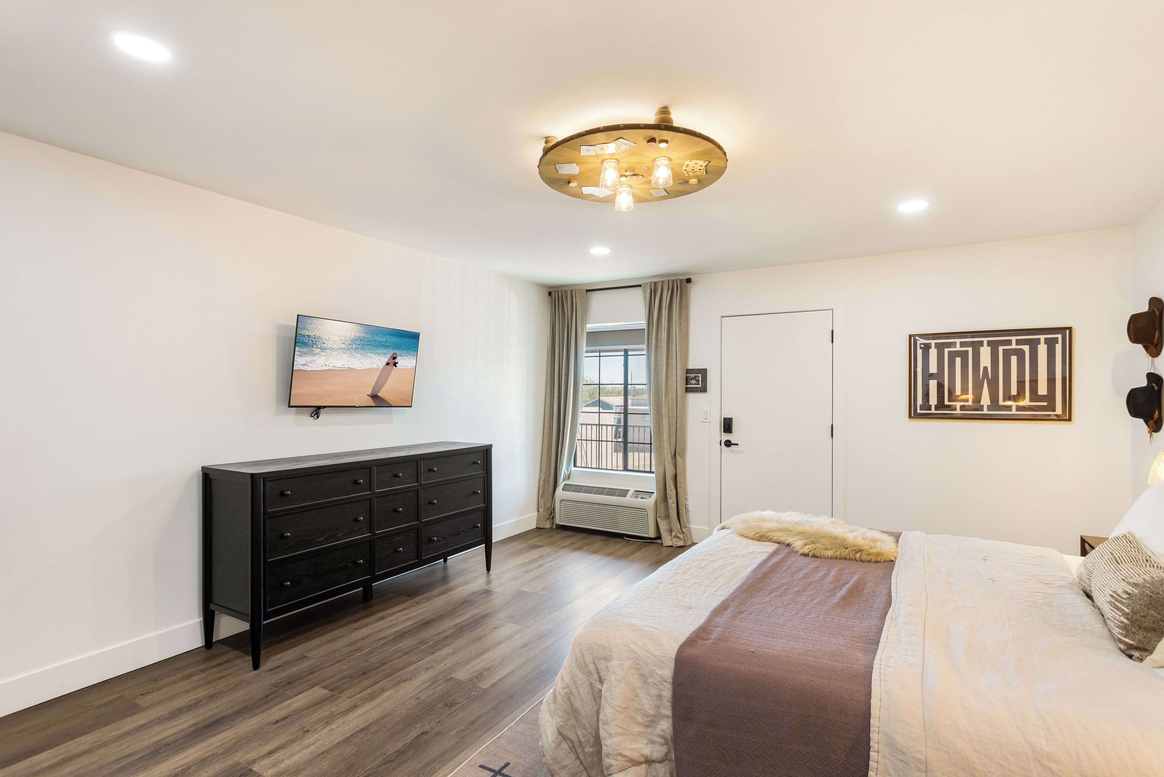 Un dormitorio con cama, cómoda, luz de techo, ventana con cortinas y una obra de arte colgada en la pared.