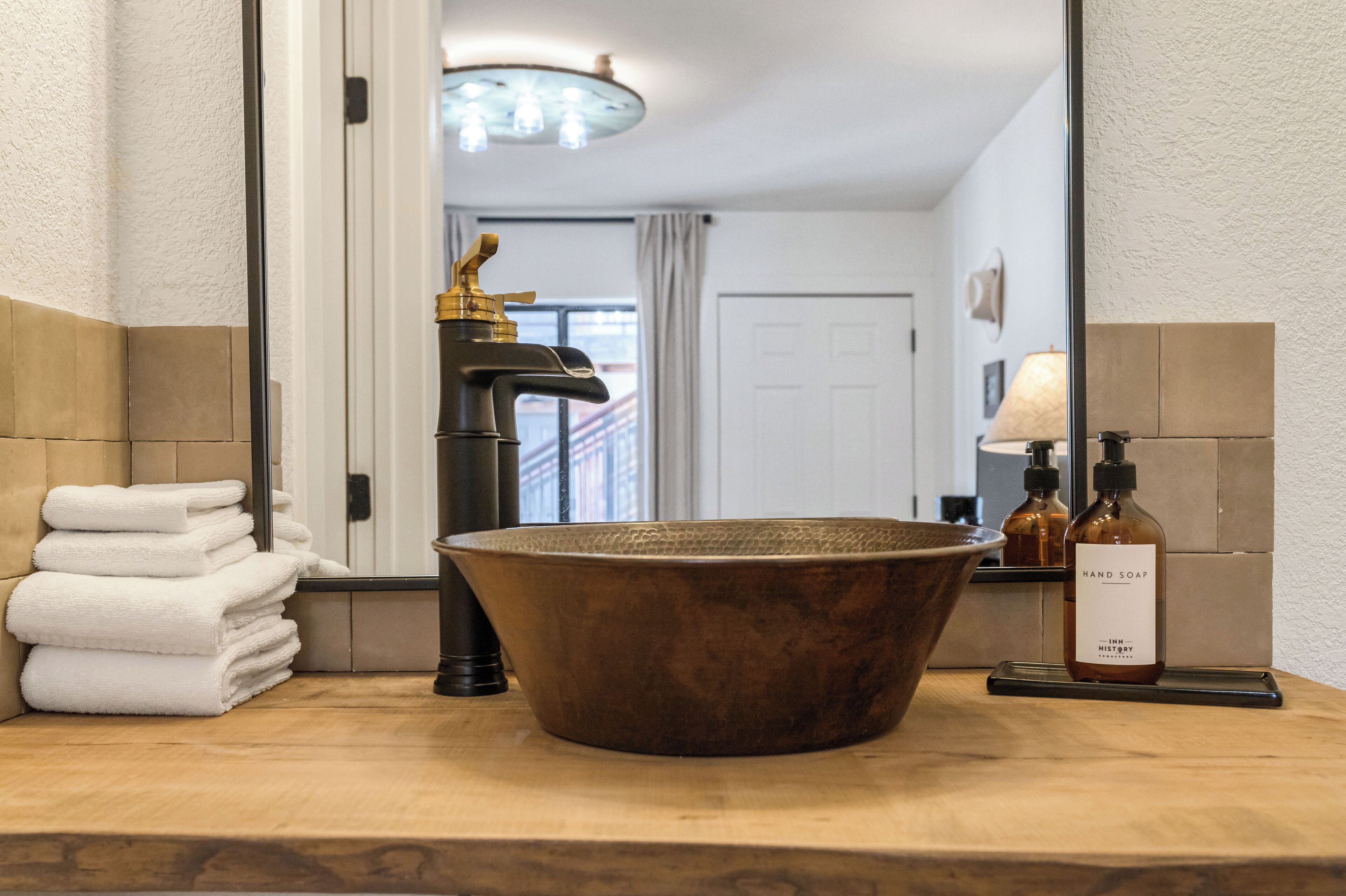 A bathroom with a large copper sink, a mirror, a stack of white towels, and a bottle of hand soap.