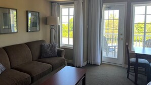 Habitación de hotel con un sofá marrón, una mesita pequeña, una lámpara de pie y vistas al exterior a través de amplias ventanas.
