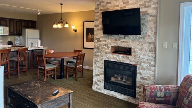 A living room with a fireplace, a television mounted on the wall, a dining area with chairs, and a kitchen in the background.