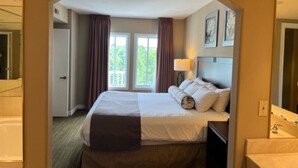 A hotel room with a bed, a nightstand, a lamp, a mirror, and a view of the outdoors through a window.