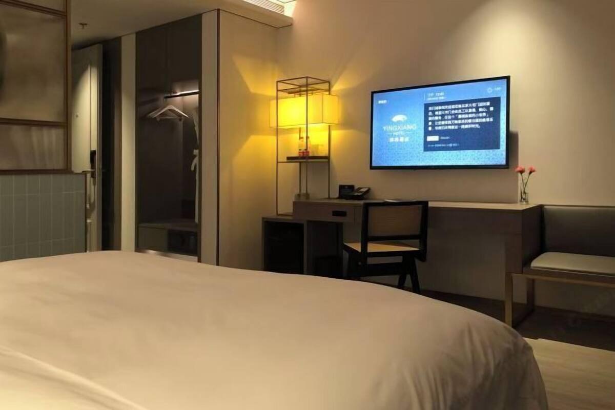A hotel room with a bed, a desk, a chair, a TV screen, a lamp, and a shower area.