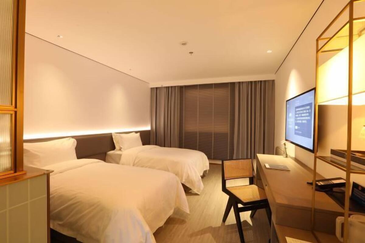 A hotel room with two beds, a desk, a chair, a TV, and a window with curtains.
