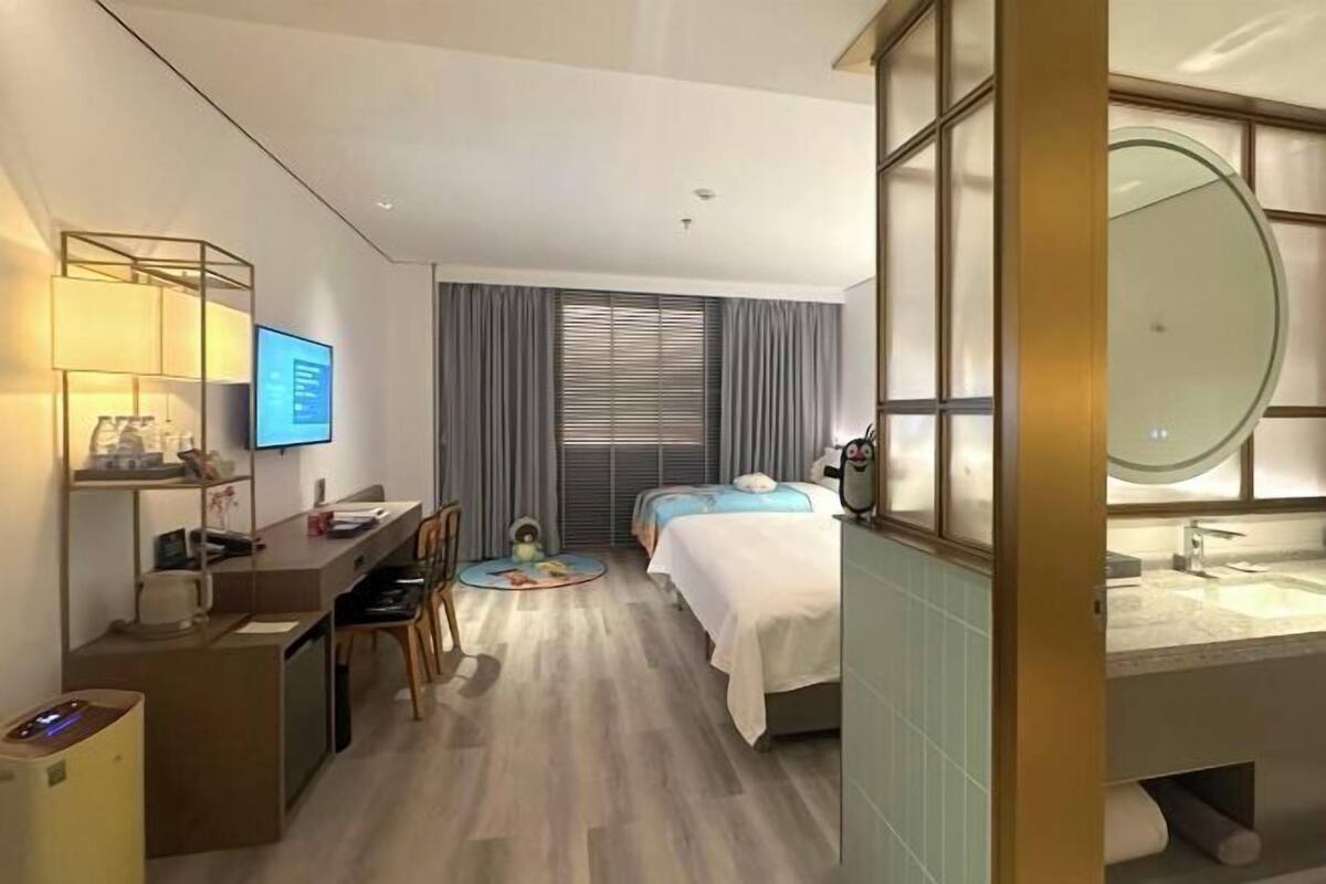 A hotel room with a desk, computer, chair, two beds, a bathroom with a large mirror, and a window with blinds.
