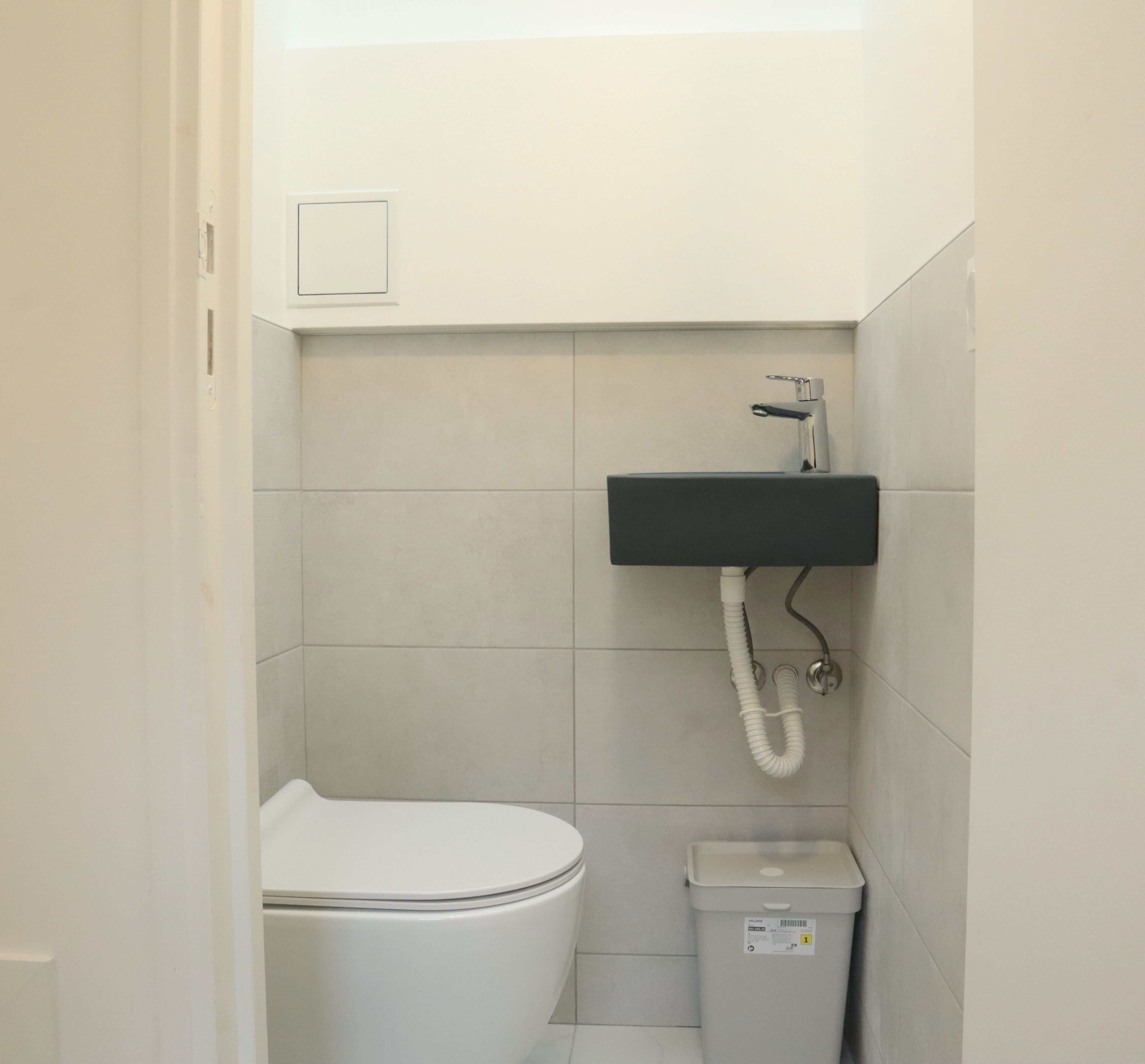 A small, clean bathroom with a toilet, a sink with a hose, and a trash bin.