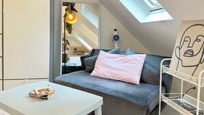 A cozy loft bedroom with a sloped ceiling, a skylight, a grey sofa with pillows, a white side table, and a metal shelf unit. - Lovely Rooftop designed Apt 200m to Panthéon  (Paris)