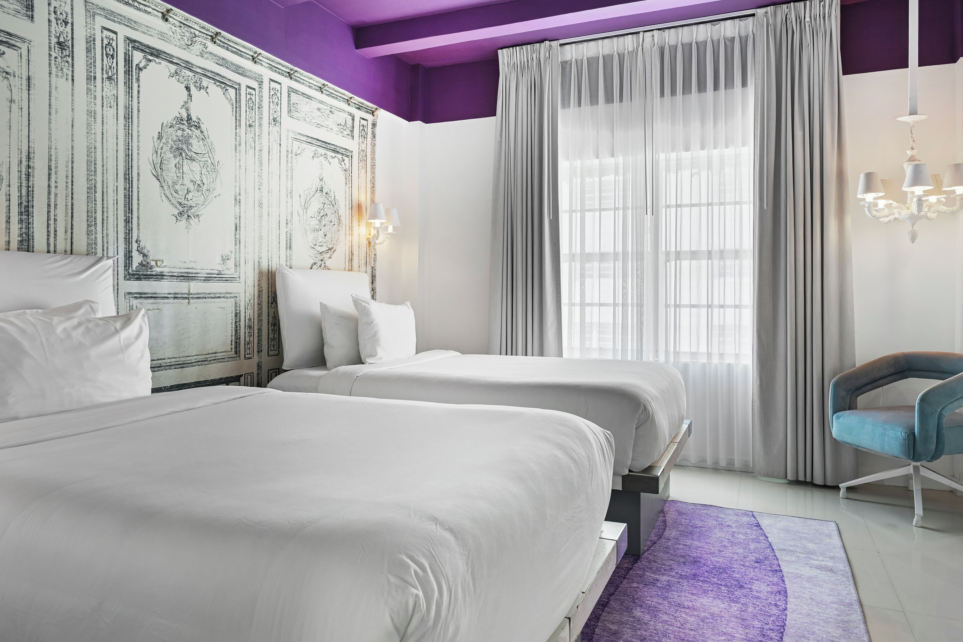 A hotel room with two beds, a purple accent wall, and a chandelier.