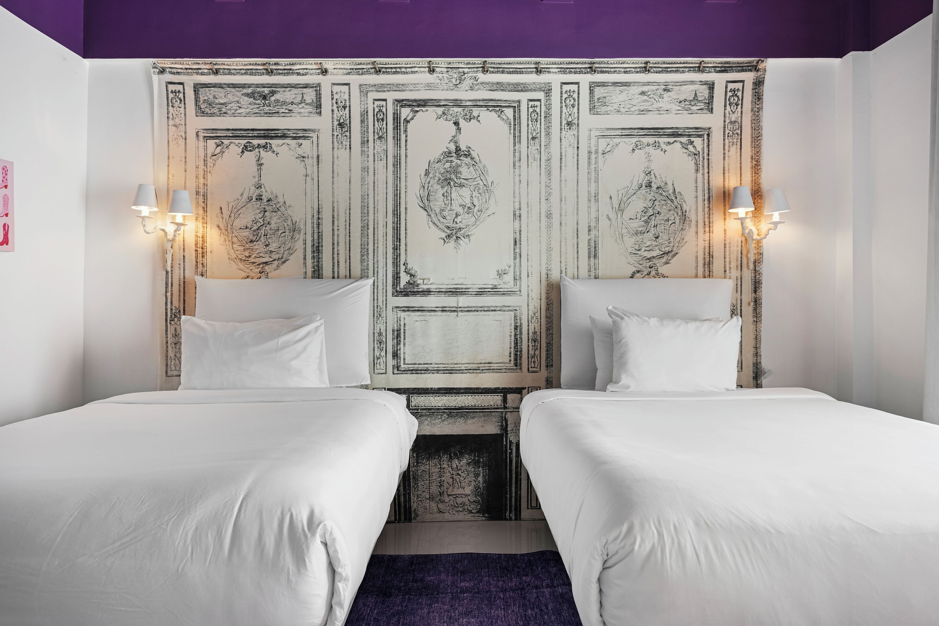 Ein Hotelzimmer mit zwei Betten, einem dekorativen Kopfteil, Wandleuchten und einem violetten Teppich.