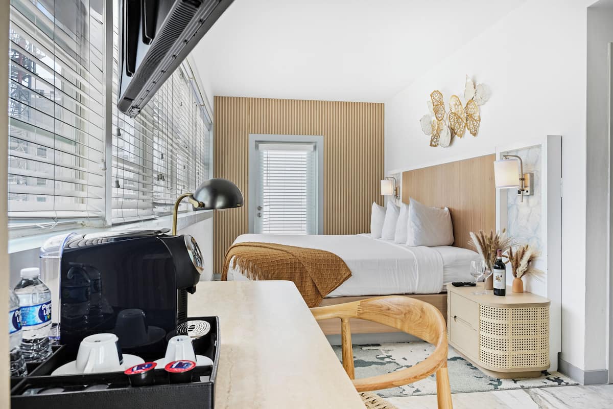 Hotellihuone, jossa on sänky, yöpöytä, lamppu ja ikkuna, jossa on sälekaihtimet.