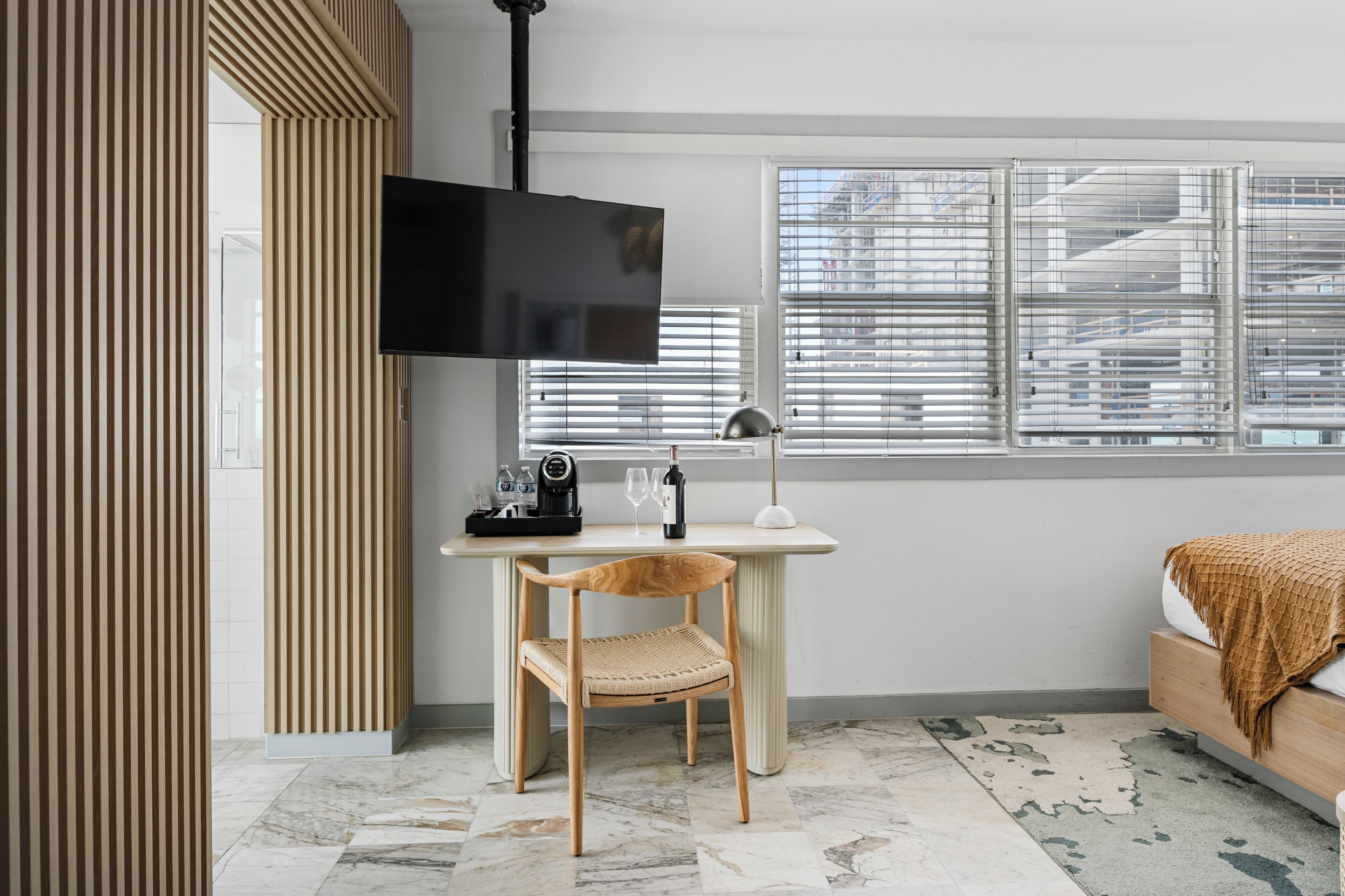 A modern room with a wooden desk, a chair, a bed, a TV, and a window with blinds.