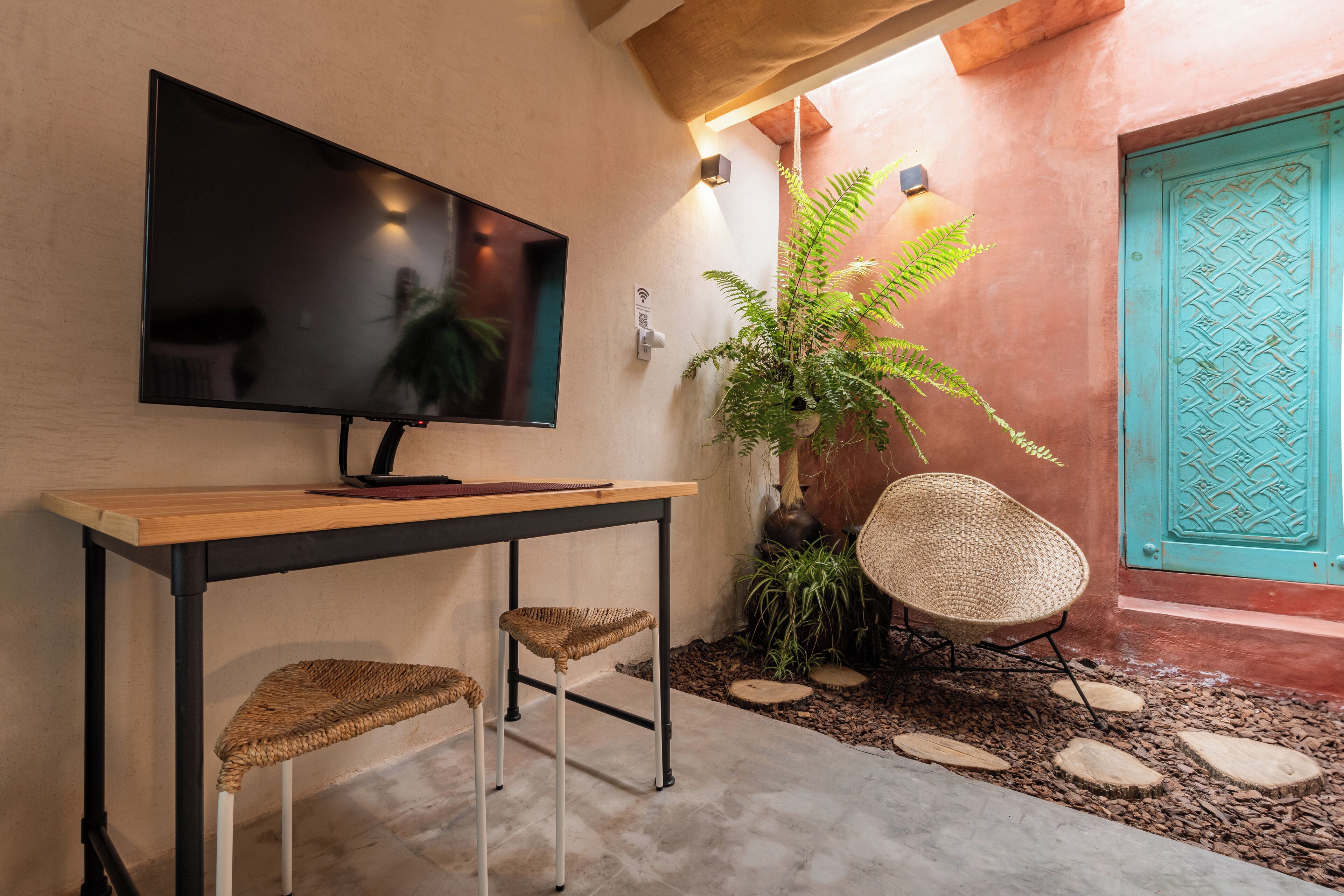 Ein modernes Interieur mit einem an der Wand befestigten Flachbildfernseher, einem Holztisch mit zwei gewebten Stühlen, einer Topfpflanze und einer türkisblauen Tür.