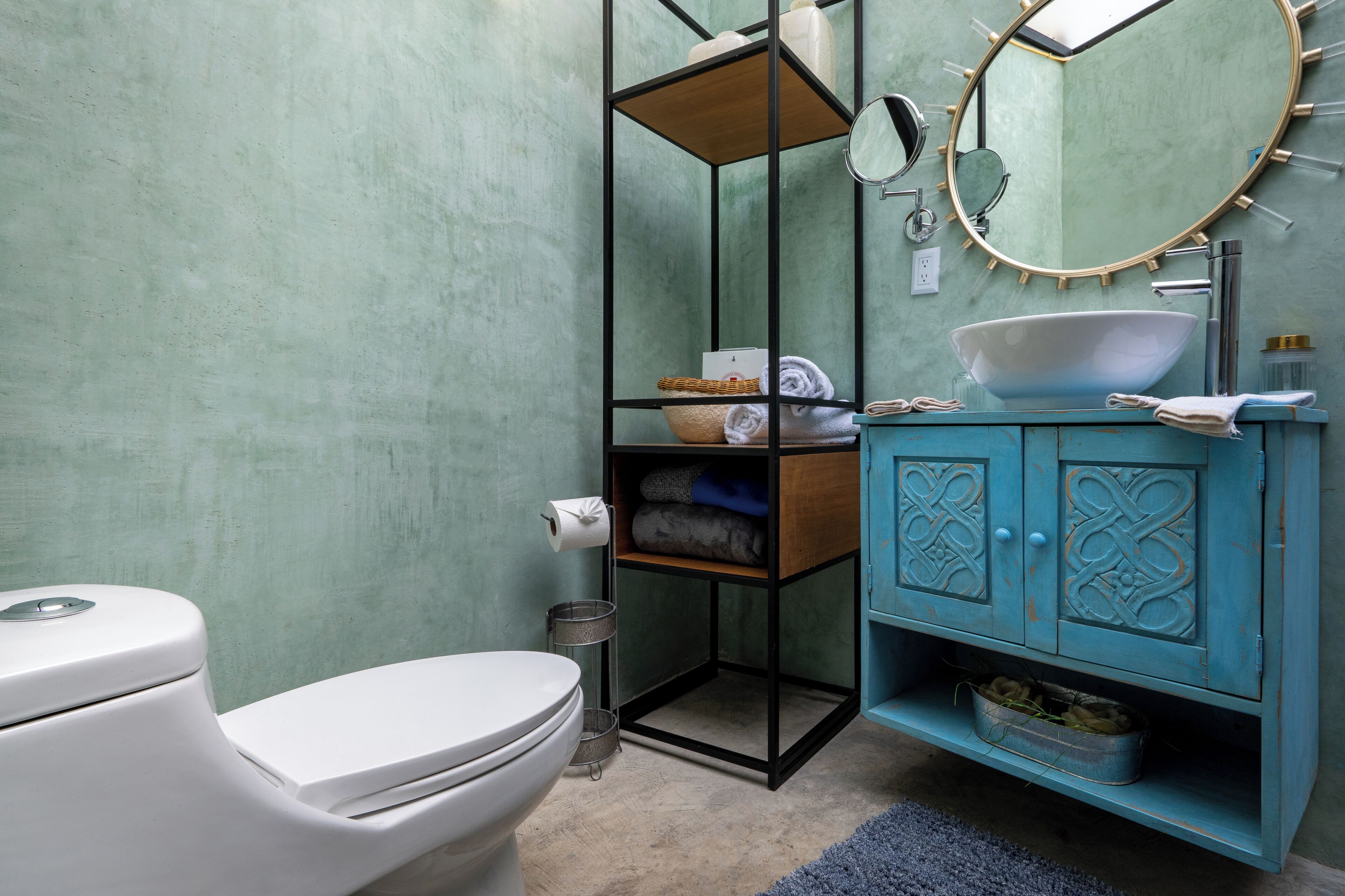 A bathroom with a white toilet, a sink with a white basin, a blue cabinet with decorative doors, a round mirror, and a shelf with towels and toiletries.