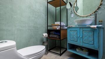 A bathroom with a white toilet, a sink with a white basin, a blue cabinet with decorative doors, a round mirror, and a shelf with towels and toiletries.