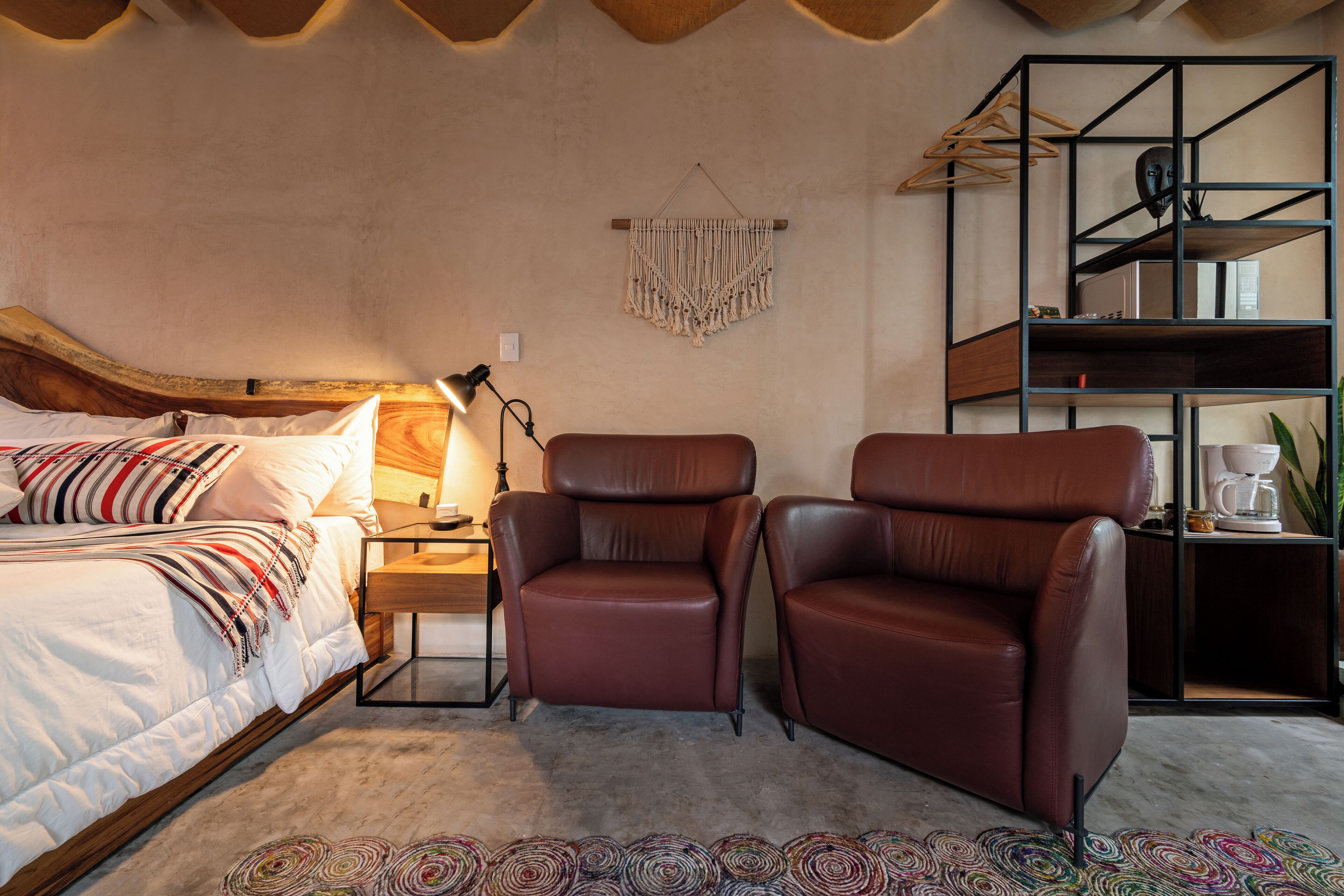 Een slaapkamer met een bed, twee leren fauteuils, een nachtkastje, een lamp, een macramé wanddecoratie en een inbouwkast met een koffiezetapparaat.