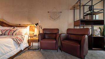 Een slaapkamer met een bed, twee leren fauteuils, een nachtkastje, een lamp, een macramé wanddecoratie en een inbouwkast met een koffiezetapparaat.