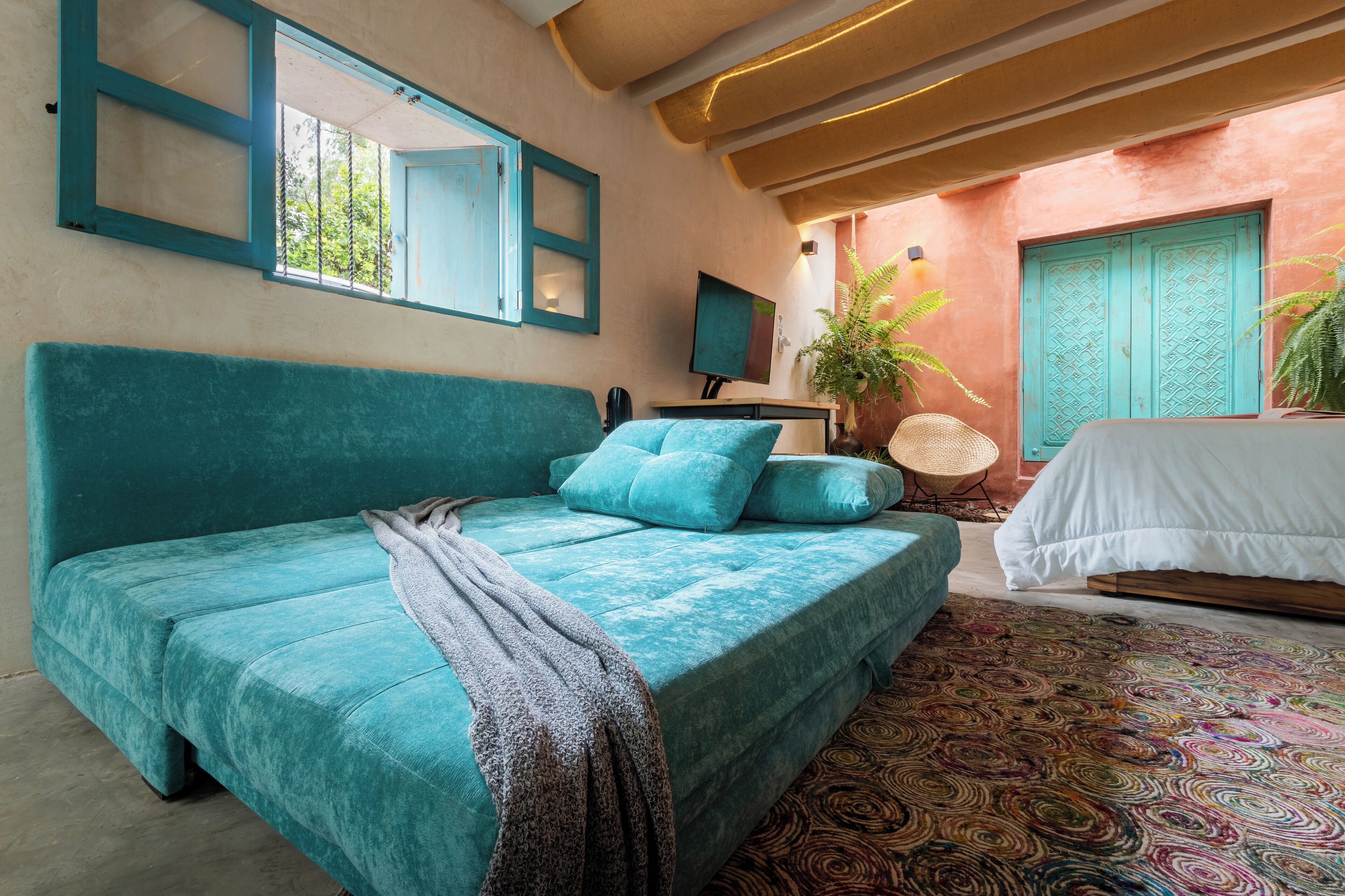 Une chambre avec un lit de couleur turquoise, un tapis à motifs, une plante en pot et une vue sur l’extérieur.