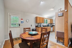 Un coin repas avec une table ronde, des chaises et un vase décoratif. On y trouve une porte avec une fenêtre, des armoires en bois et un petit réfrigérateur.