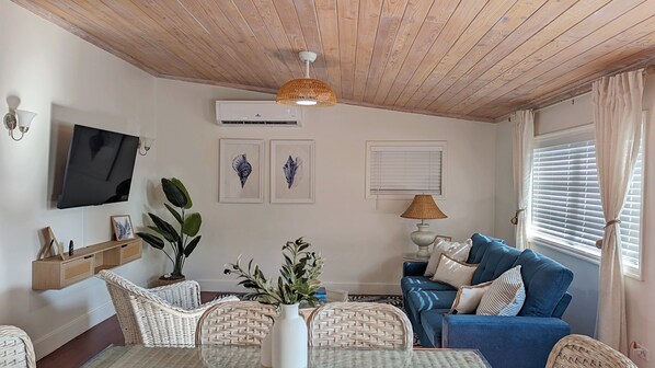 Un salon avec un plafond en bois, un téléviseur à écran plat fixé au mur, un canapé bleu avec des coussins à rayures, des meubles en osier et des œuvres d’art encadrées accrochées au mur.