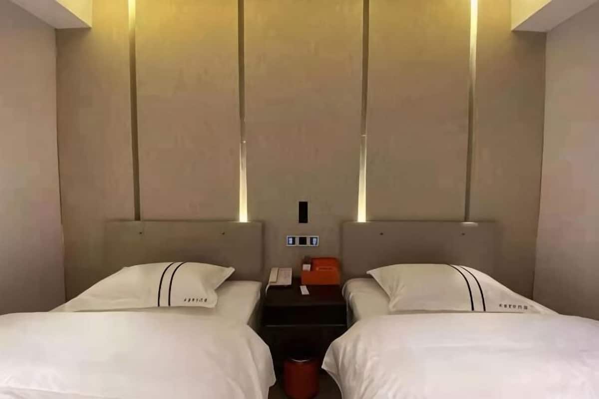 Habitación de hotel con dos camas, una mesita de noche, un cesto de basura rojo y un panel de control en la pared.