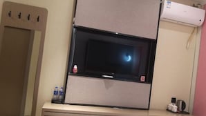 Ein Hotelzimmer mit einem an der Wand befestigten Flachbildfernseher, einem kleinen Tisch mit einer Wasserflasche, einer Tasse und einer Fernbedienung.