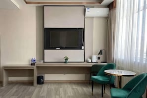 Una habitación de hotel con un televisor de pantalla plana grande montado en la pared, una mesita con una silla, una planta en maceta y una ventana con cortinas.