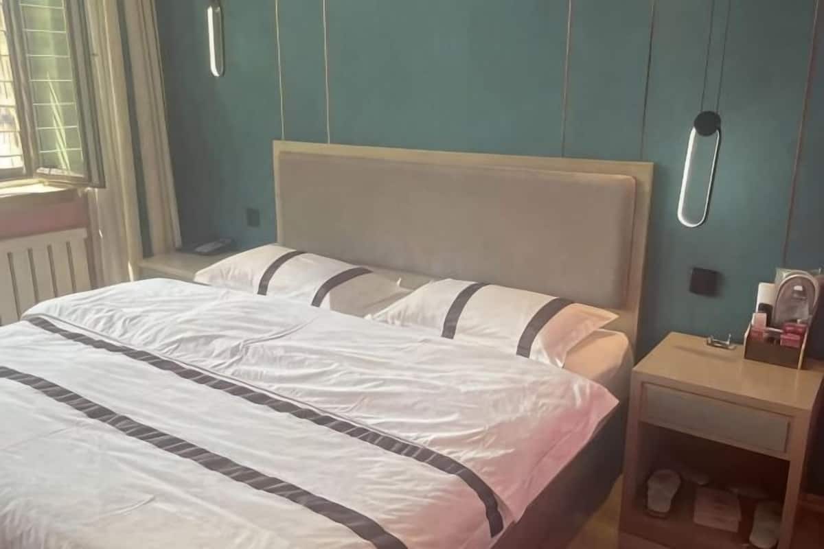 Una cama con ropa blanca y un cabecero de madera, una mesita de noche con una lámpara, y una ventana con cortinas.