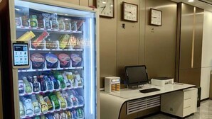Ein Getränkeautomat mit Getränken und Snacks, ein Schreibtisch mit Computer und Uhren an der Wand.