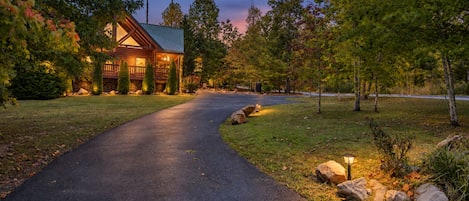 A paved driveway leading to a wooden cabin with a metal roof, surrounded by trees and a well-maintained lawn.