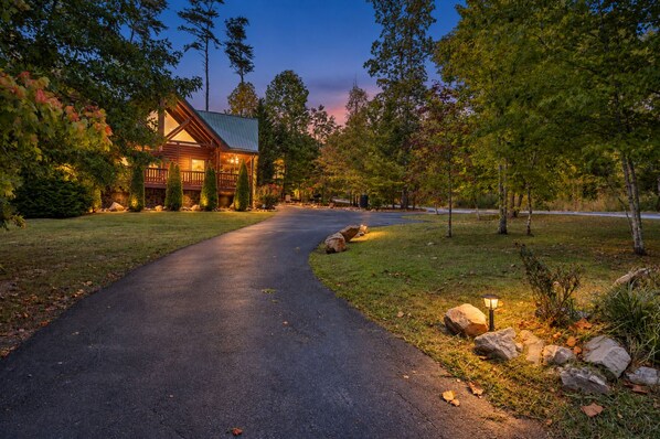 Une allée pavée mène à un chalet en bois surmonté d’un toit en métal, entouré d’arbres et bordé d’une pelouse bien entretenue.